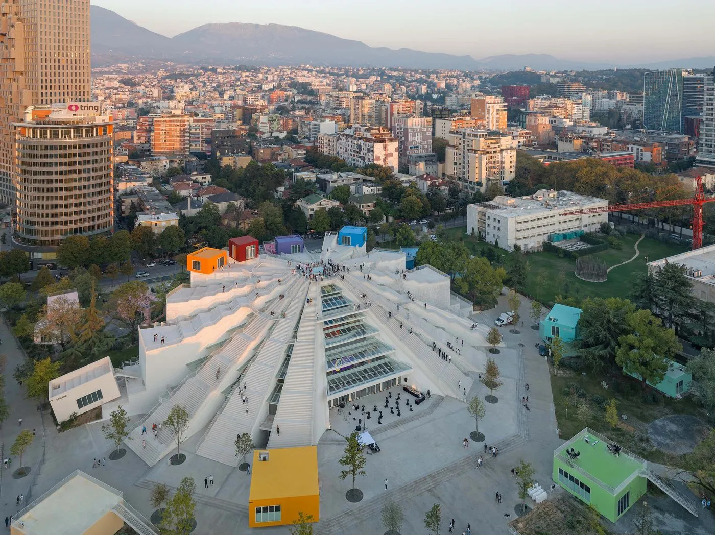 Pamje ajrore nga afër e Piramidës së Tiranës, me njerëz që ngjiten në shkallët e saj dhe kubet me ngjyra të ndezura, e vendosur në zemër të qytetit.