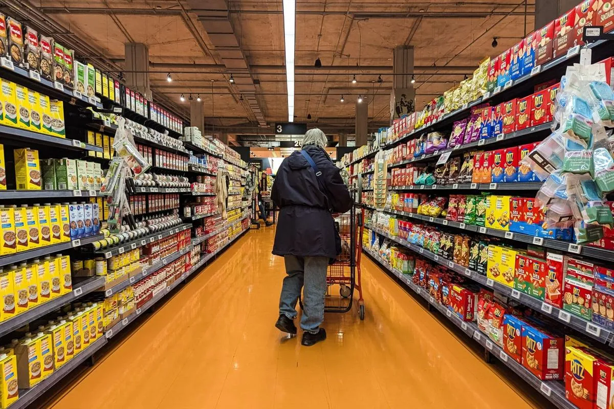 Një person ecën nëpër një korridor supermarketi, i rrethuar nga rafte të mbushura me produkte ushqimore të paketuara.