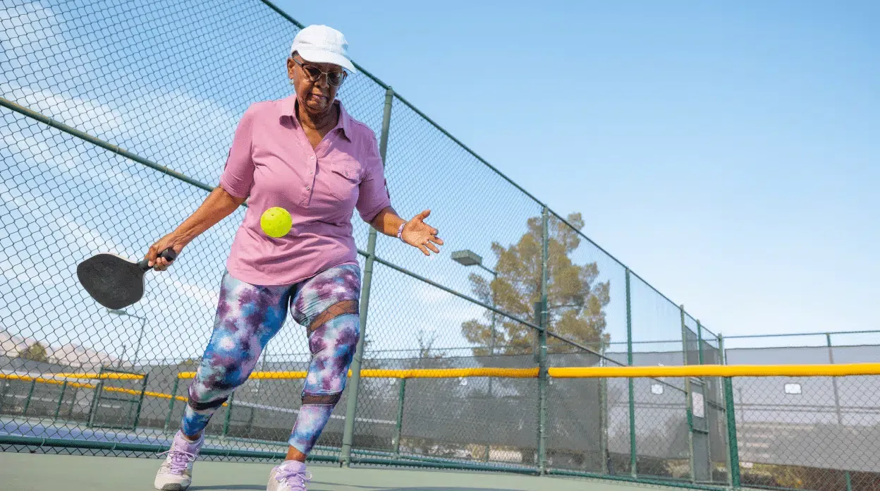Një grua e moshuar me kapelë duke luajtur pickleball, duke ilustruar rëndësinë e aktivitetit fizik për plakjen e shëndetshme.