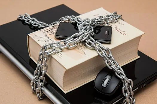 Një libër dhe dy telefona të lidhur me zinxhirë mbi një laptop, simbolizojnë mbrojtjen e të dhënave personale dhe sigurinë e pajisjeve dixhitale.